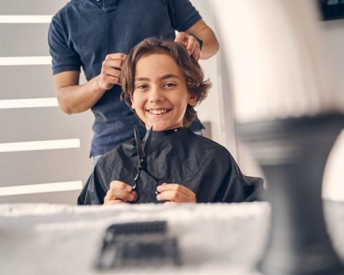 Caucasian child sitting quietly on chair while hairdresser in casual clothes doing him haircut at home